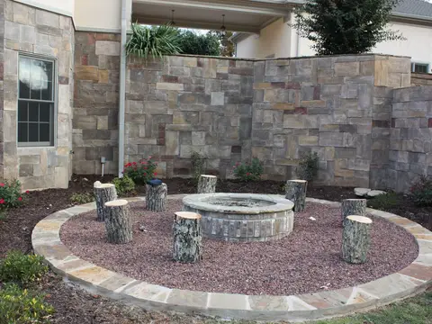 Circular stone fire pit with log-stump seats and a stone-edged gravel ring in front of a stone wall