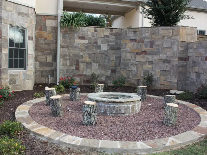Circular stone fire pit with log-stump seats and a stone-edged gravel ring in front of a stone wall