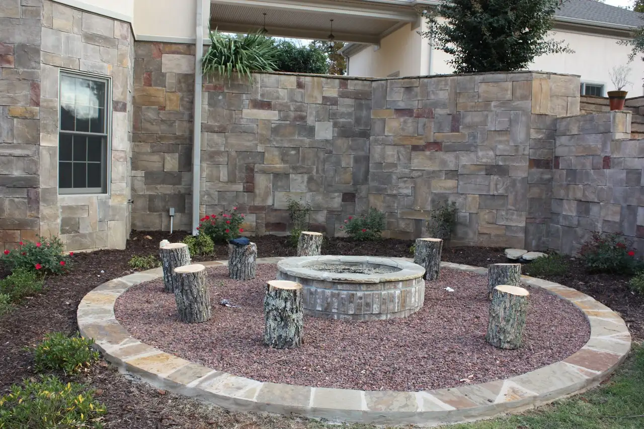 Circular stone fire pit with log-stump seats and a stone-edged gravel ring in front of a stone wall