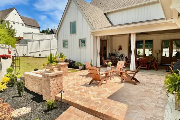 Herringbone brick paver patio with Adirondack chairs beside the covered porch of a white farmhouse-style home